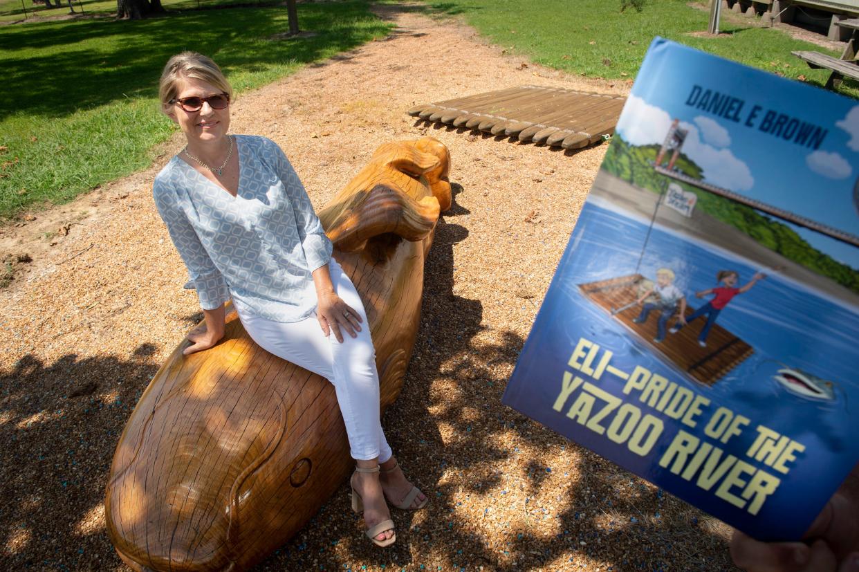 Michele Harris, the event coordinator for the Eli — Pride of the Yazoo River Festival, sits on the Eli wooden sculpture on Plum Street in Satartia, Miss., Tuesday, July 19, 2022. The festival, July 30, 2022,  celebrates the fictitious giant catfish, Eli, who is the subject of a children's book written by Daniel Brown, “Eli — Pride of the Yazoo River,” right. In the book, Eli lives under the Yazoo River bridge in Satartia.