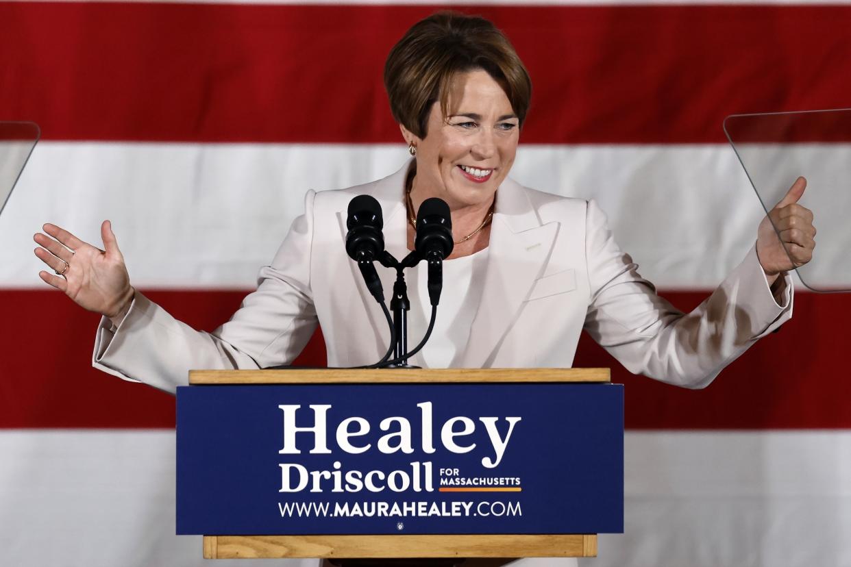 Maura Healey, wearing a white suit, gives a thumbs-up while standing at a lectern.