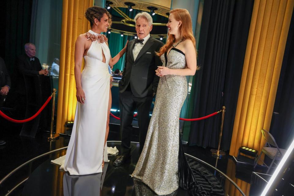 Two women in evening gowns flank a man in a tux backstage at the Oscars.