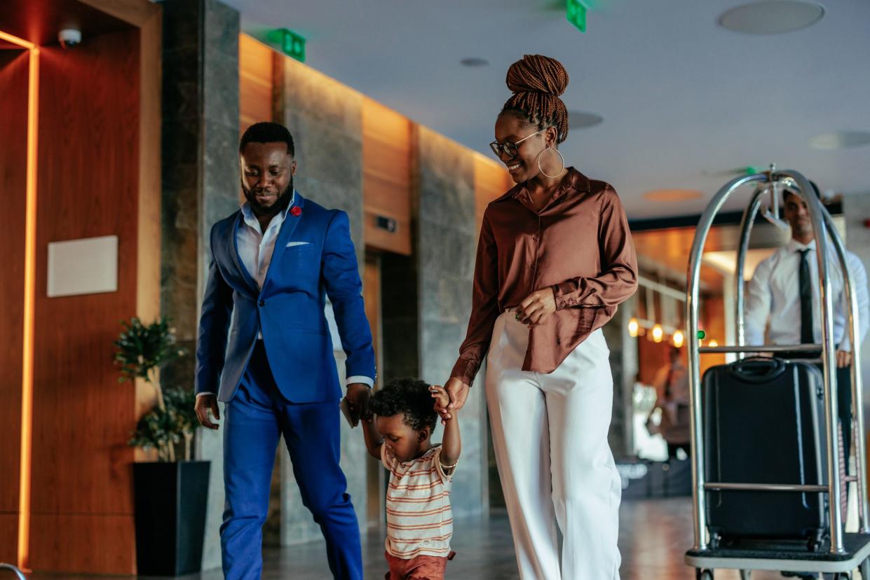 An elegant African American family with their child is in a luxurious hotel, walking through a lobby with the hotel attendant behind them with their luggage.