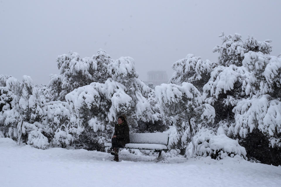 A woman sits on a snow-covered bench in front of the Parthenon temple, in Athens, Tuesday, Feb. 16, 2021. Unusually heavy snowfall has blanketed central Athens, with authorities warning residents particularly in the Greek capital's northern and eastern suburbs to avoid leaving their homes. (AP Photo/Petros Giannakouris)