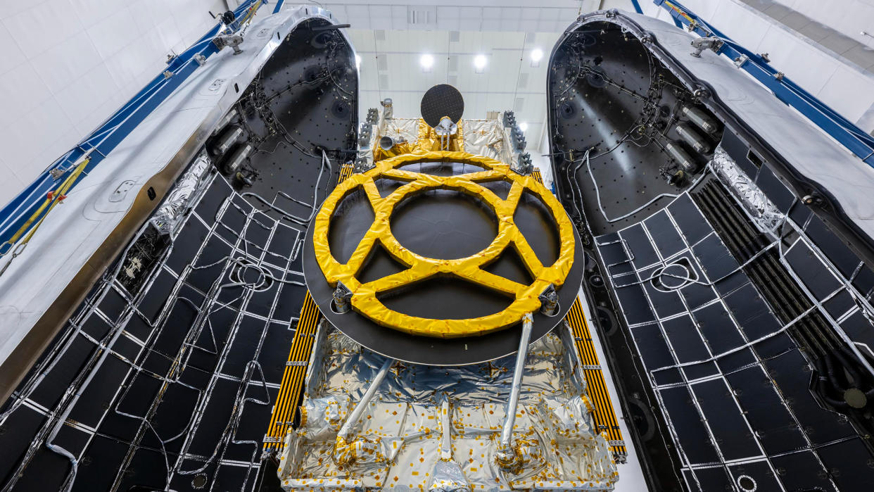  A black and yellow satellite is seen between the two halves of its rocket's payload fairing inside a white-walled building. 