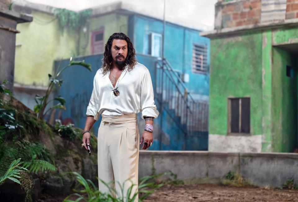 Jason Momoa in a white shirt and cream pants with jewelry, on a tropical set