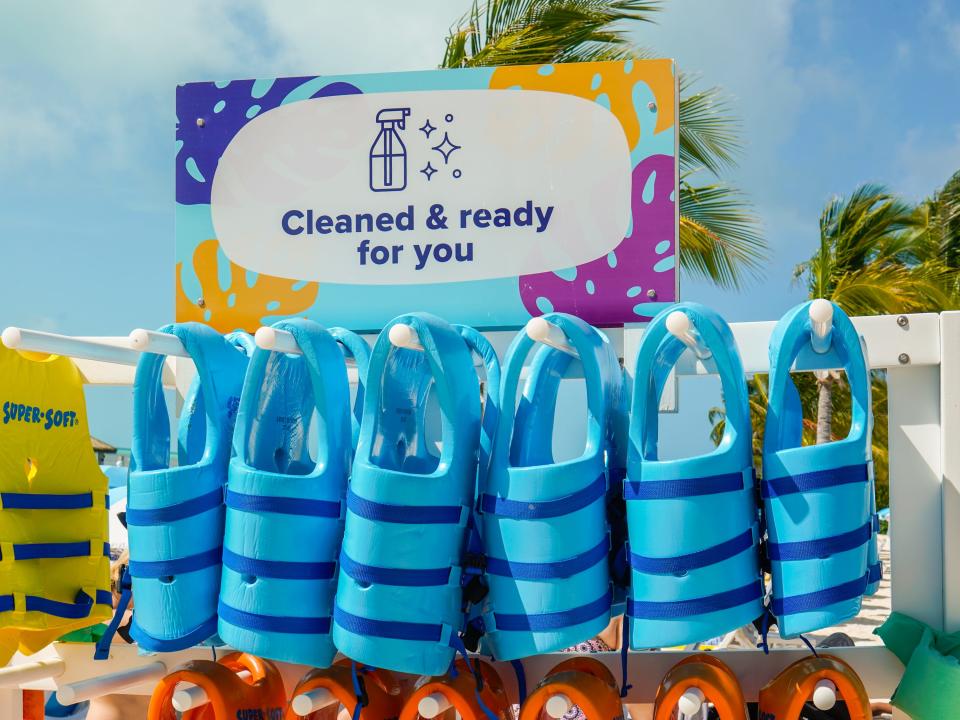 A sign says "Cleaned and ready for you" above a rack of blue life vests on the beach at CocoCay with palm trees and cloudy blue skies in the background