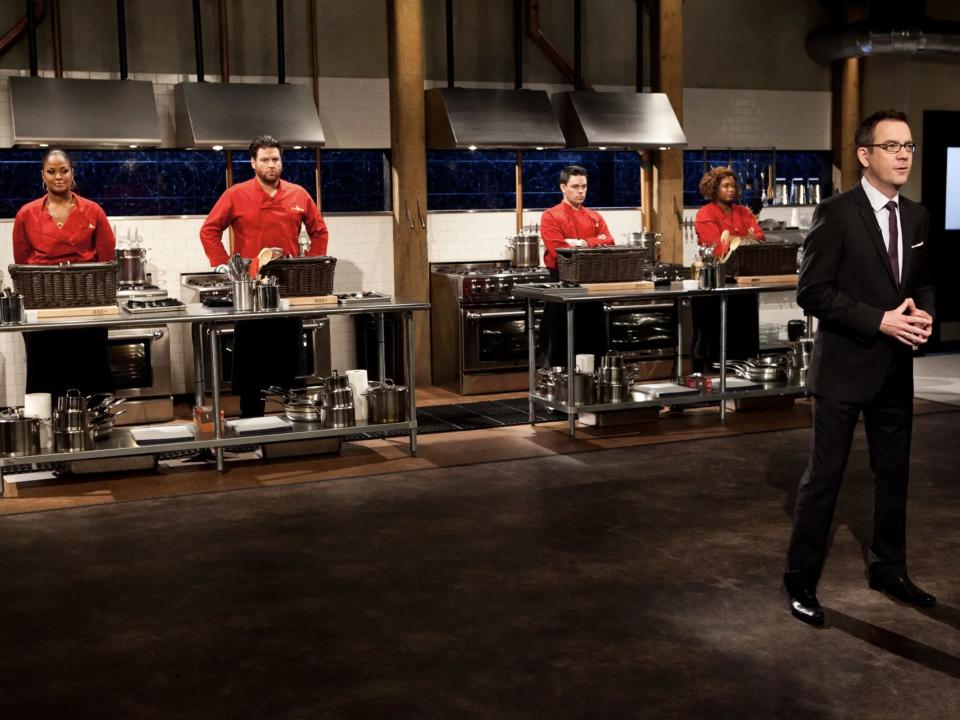 Chefs in front of cooking stations with unopened baskets in front of them with Ted Allen in front on set of "Chopped"
