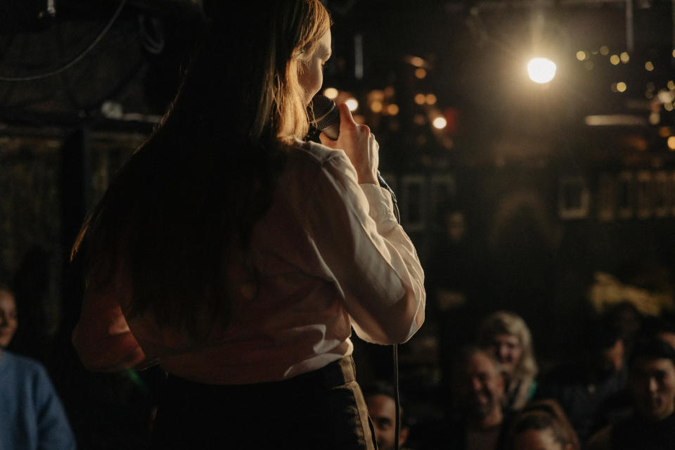 Person holding a microphone, speaking to an audience in a dimly lit indoor setting