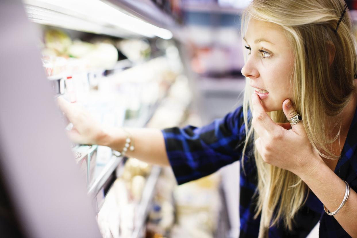 woman checks the supermarket fridge's contents, hand to her mouth as she chooses