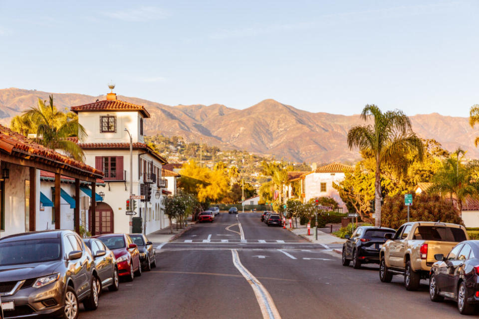 Pictured: Cars lined up on both sides of a street filled with houses and a mountain in the background.