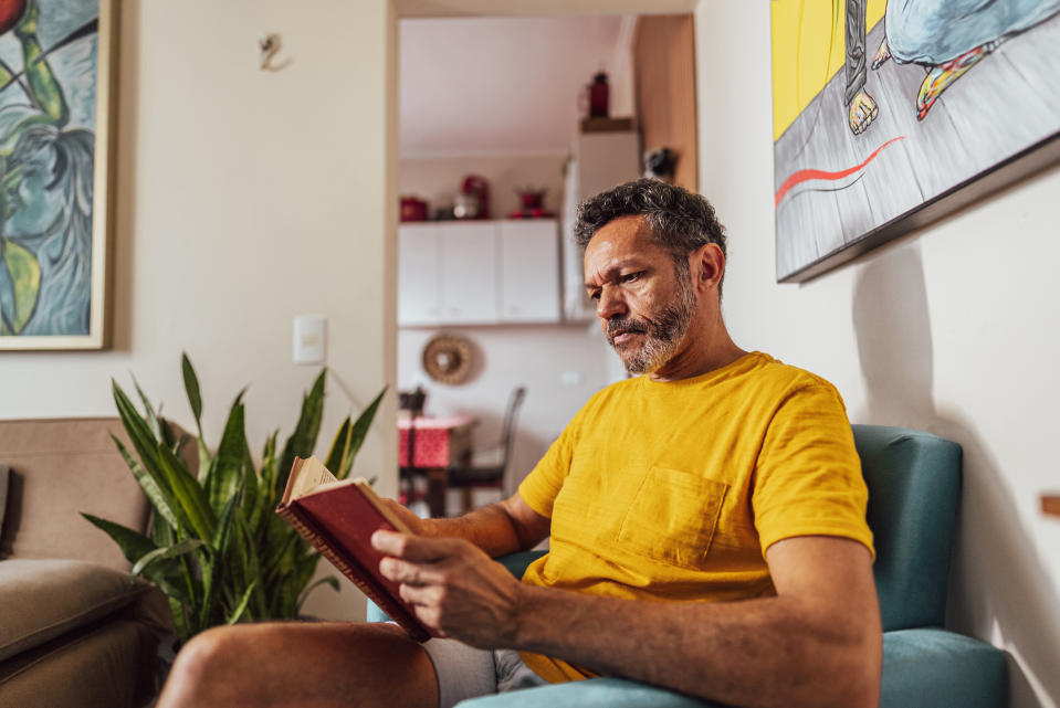 A man with gray hair wearing a yellow shirt sits in a cozy living room, reading a book. There are plants and artworks in the background