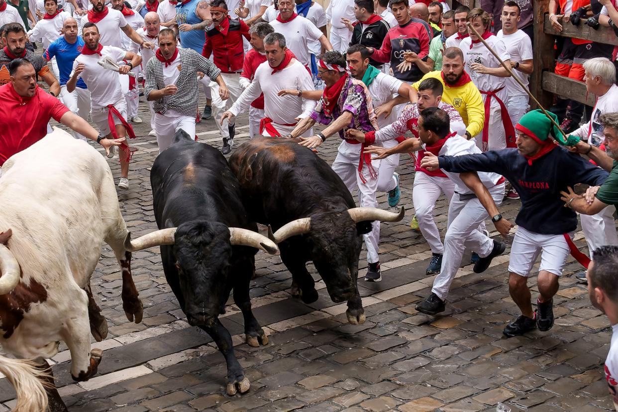 Participants, many wearing all white and red scarves, run alongside three bulls on a cobblestone street.