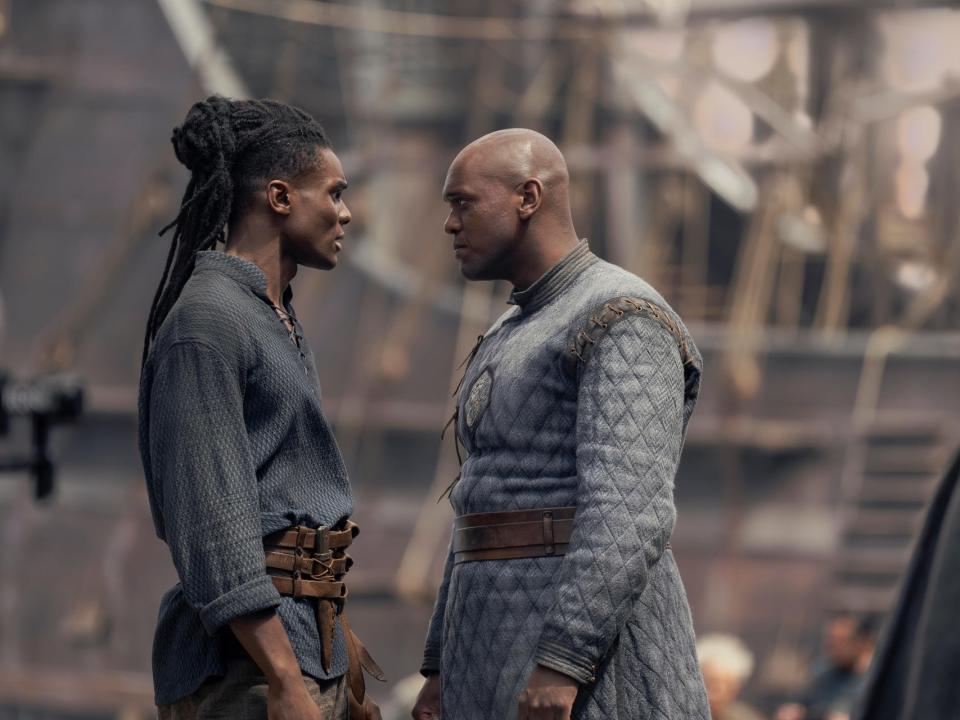 clinton liberty and abubakar salim as addam and alyn of hull, two young men in blue clothing. addam has long hair, arranged in dreadlocks and pulled back, while alyn is bad. they're looking at each other in a shipyard