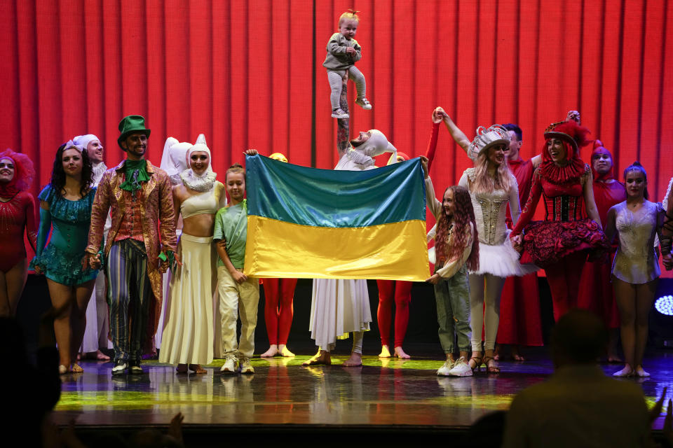 Performers stand on stage with the Ukrainian flag, held by two of their children, and baby Milena hoisted up above it at the end of the show "Alice in Wonderland" in Pistoia, Italy, Friday, May 6, 2022. A Ukrainian circus troupe is performing a never-ending “Alice in Wonderland” tour of Italy. They are caught in the real-world rabbit hole of having to create joyful performances on stage while their families at home are living through war. The tour of the Theatre Circus Elysium of Kyiv was originally scheduled to end in mid-March. (AP Photo/Alessandra Tarantino)