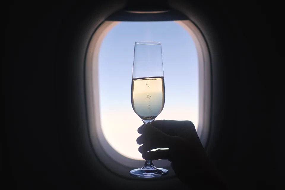 A hand is holding a champagne glass up to an airplane window, with the sky visible outside