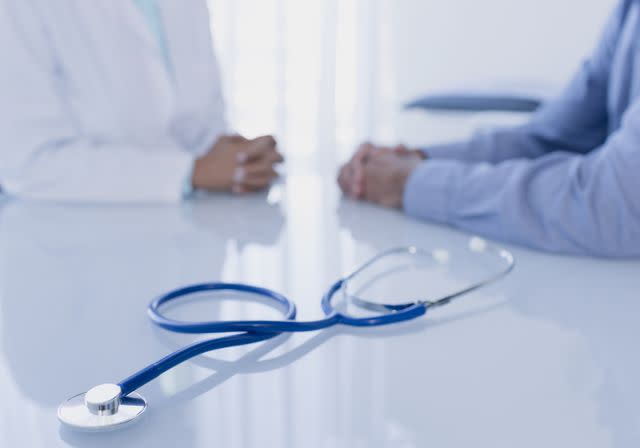 Dan Dalton / Getty Images Stock image of stethoscope on white desk in doctors office, female doctor and patient sitting in background