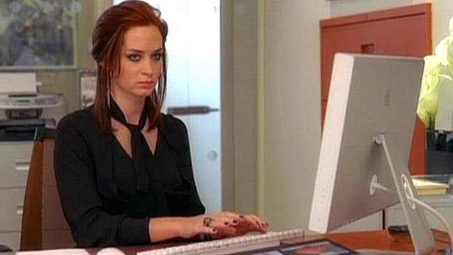 Woman at desk looking at a computer screen, wearing a black v-neck top with her hair down