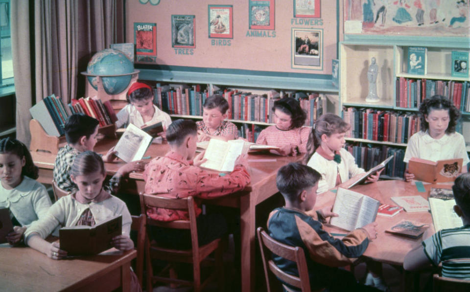 Students sitting at desks and reading books in a classroom lined with bookshelves, a globe, and educational posters
