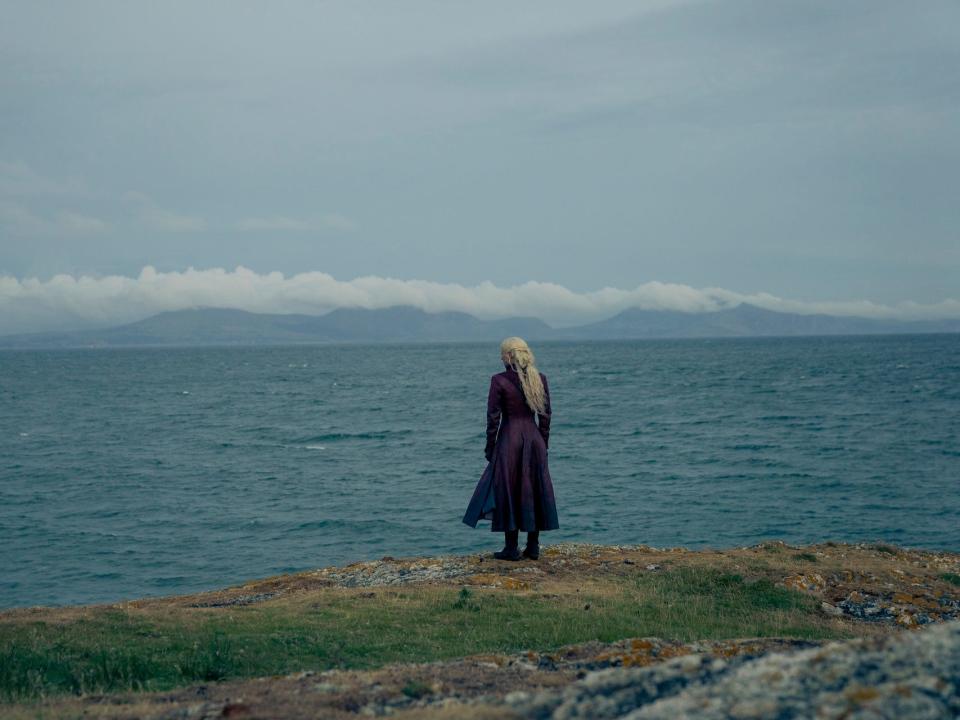 emma d'arcy as rhaenyra targaryen, seen from the back. rhaenyra's wearing a red lather coat, her hair loose, and she's seen from the back as she looks out to the se