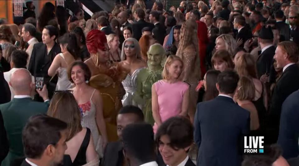 a shot of the emmys red carpet, with a crowd of people. right in the center, there's someone dressed up as a goblin, wearing a form fitting green dress