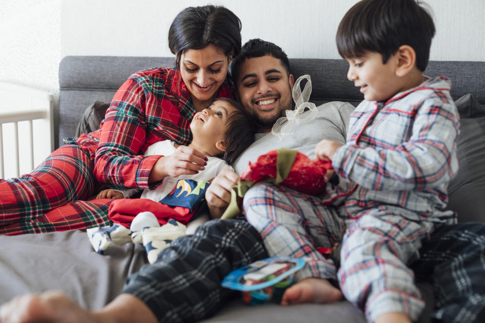 A couple sitting on their bed with their two sons during the Christmas period. The eldest son is opening a present out a stocking as his father watches. His mother is looking at his brother and smiling.