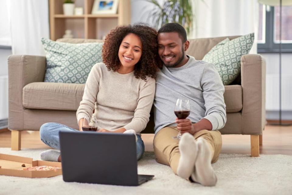 leisure, people and technology concept – happy african american couple with laptop computer and takeaway pizza and drinking red wine at home