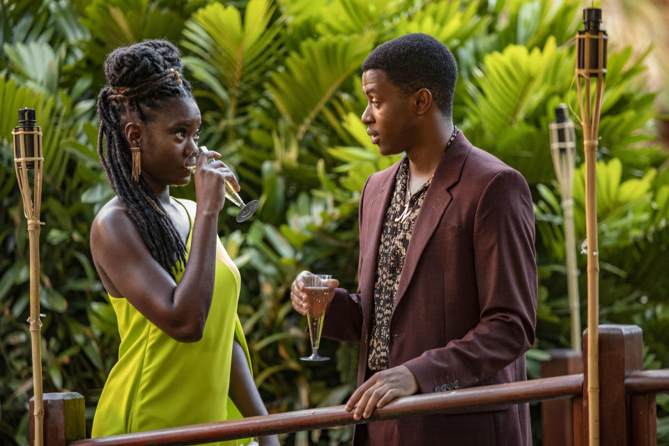 Naomi (Shantol Jackson) stands wearing a smart pale lime-coloured evening dress and sipping a glass of fizz, opposite Marlon (Tahj Miles) who is in a maroon suit and a floral shirt, also holding a glass of fizz