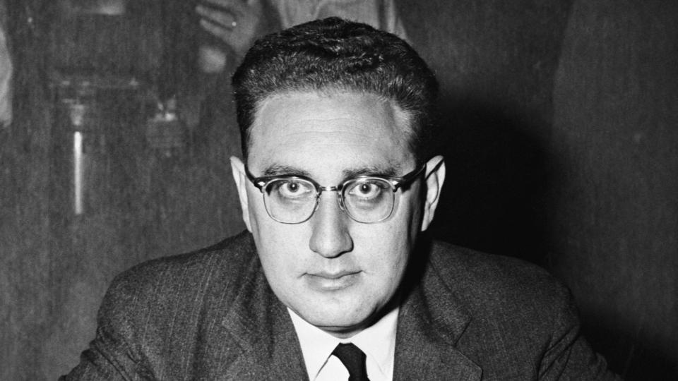 henry kissinger seated at desk looks at the camera with his hands clasped together on the table, he wears a suit jacket, tie, and collared shirt, a name plate on the desk says dr henry kissinger