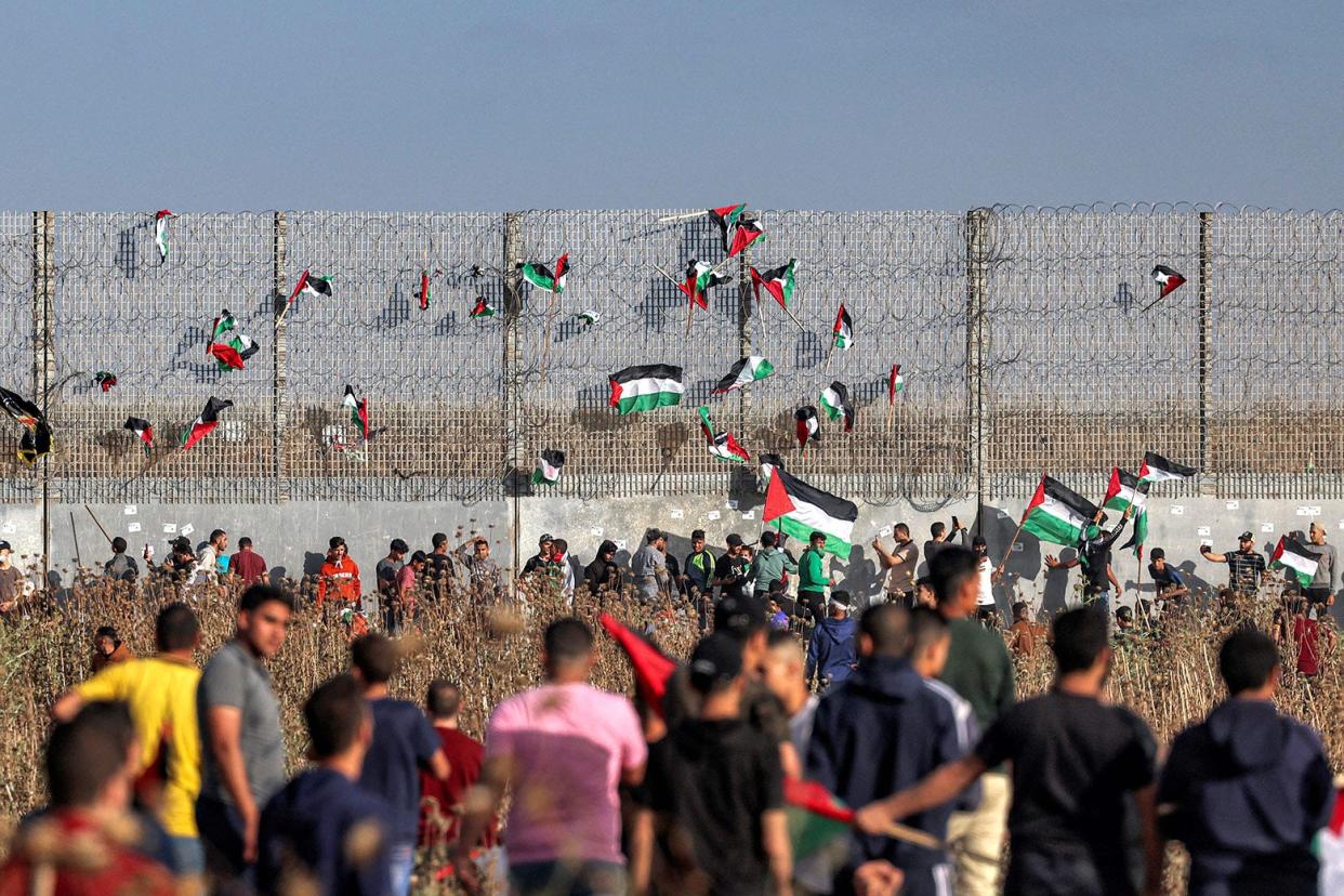 A large group of people stands in a field in front of a stretch of border fence with barbed wire. Palestinian flags are stuck in the fence.