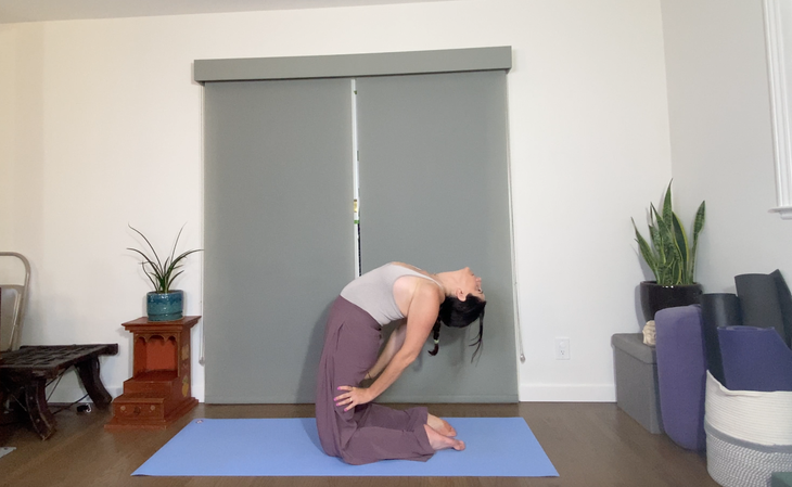 A woman in loose purple pants practices a variation of Camel Pose(Ustrasana) Laghu Vajrasana on a blue mat