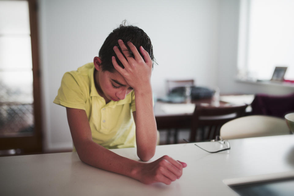 A young boy in a yellow shirt sits at a kitchen counter with his head resting on his hand, appearing deep in thought