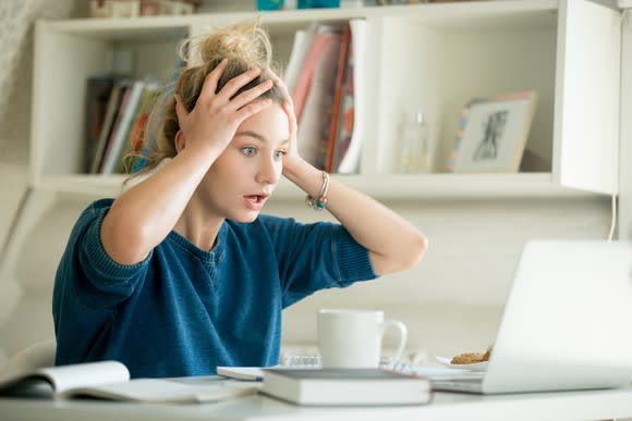 Woman at a desk grabbing her head with both hands in shock as she looks at a laptop.