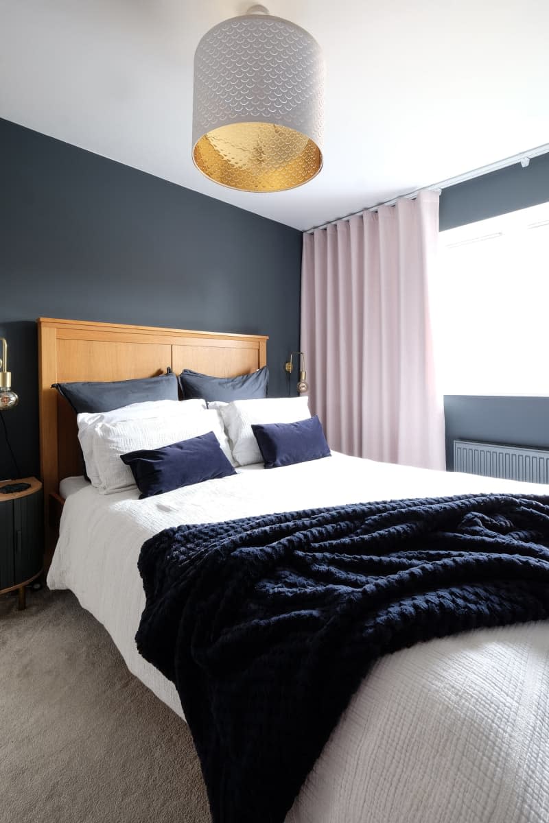 slate gray walls with light wood headboard, gold sconce fixture and white linens with dark blue accent pillows, cream curtains, large white and gold lighting fixture