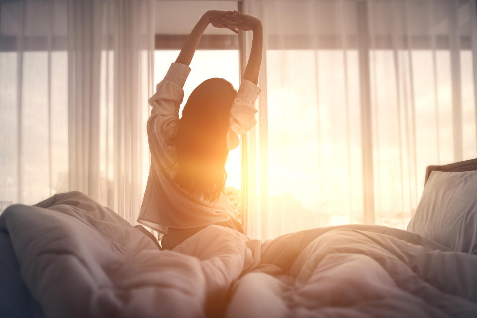 Happy woman stretching in bed after waking up, with the sun streaming in the window.