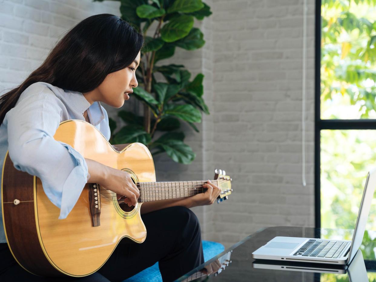 Young Asian woman practicing and learning how to play guitar on laptop computer monitor. Female guitarist watching online tutorial.