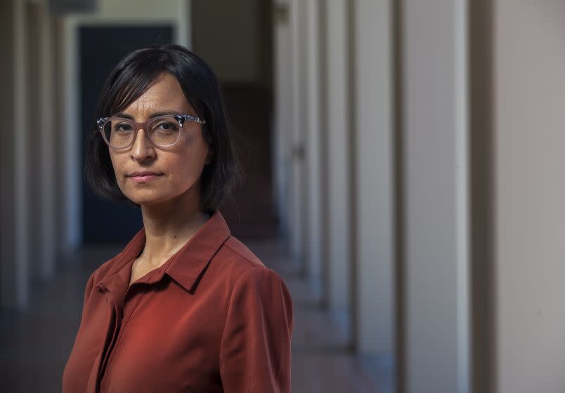 Carribean Fragoza, in a rust colored shirt and glasses, stands before a Modernist colonnade