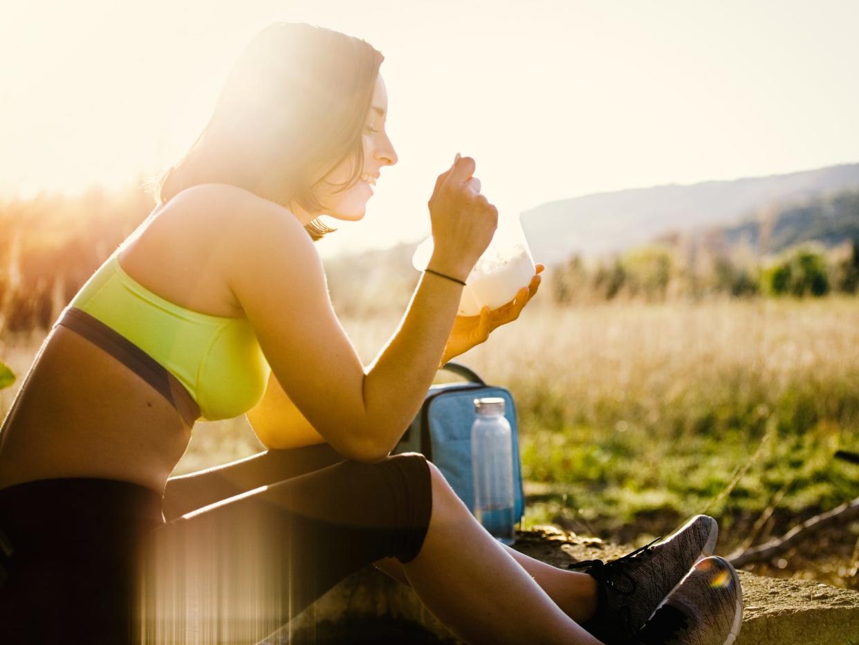 a woman in athletic clothes eating a snack on an outdoor run surrounding by a grassy landscape