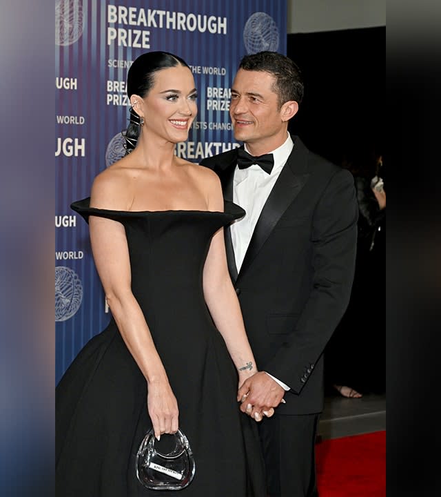 Katy Perry in a black off the shoulder gown holds hands with Orlando Bloom in a tuxedo who lovingly looks at her