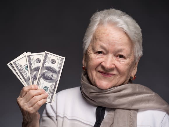 A senior woman holding up a few fanned hundred dollar bills in her right hand.