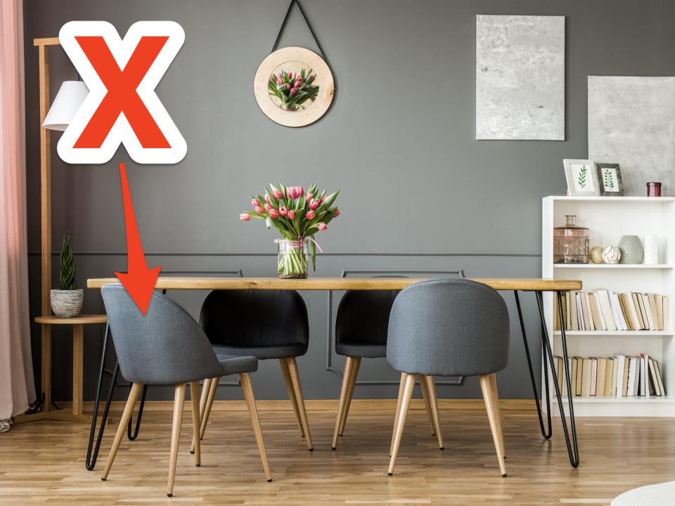 red x and arrow over photo of  modern style dining room with wooden table and gray fabric chairs with rounded low backs with bookshelves in background