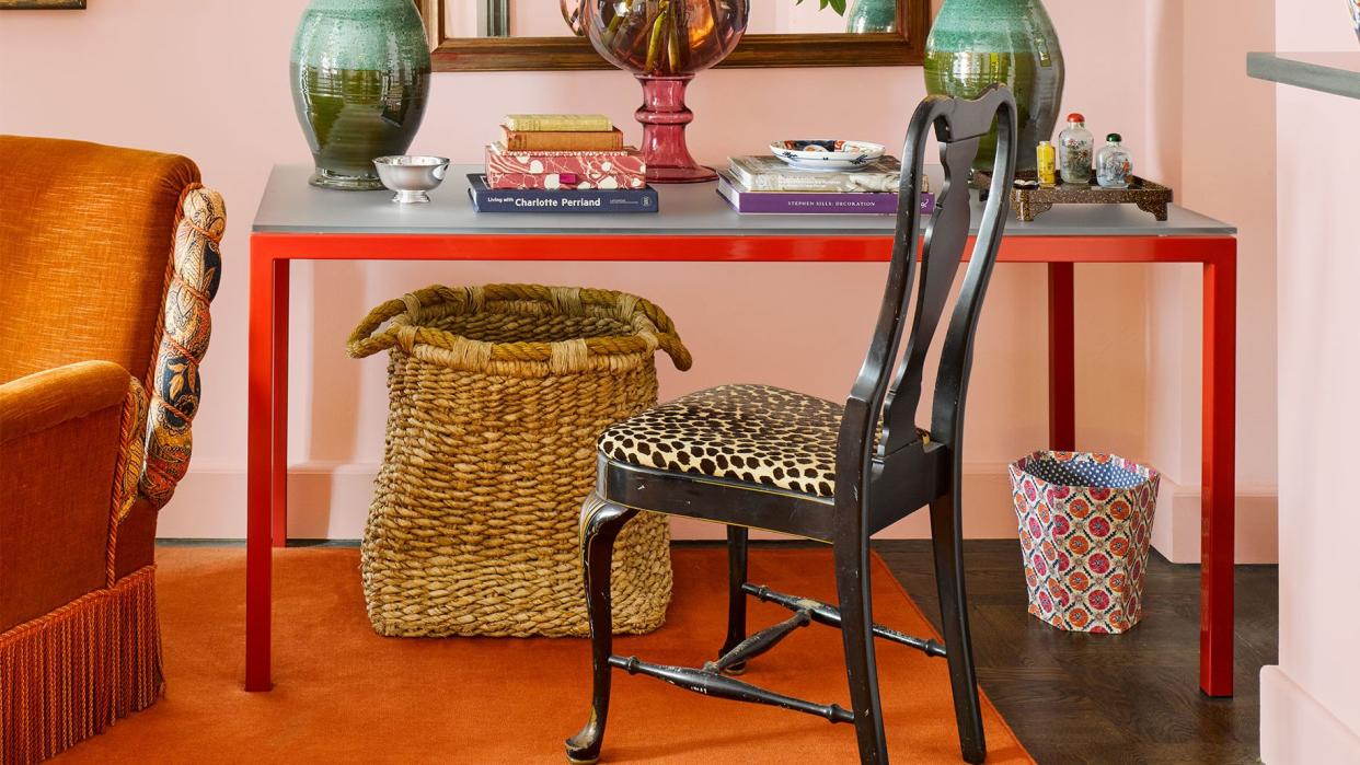 in a corner with pale pink walls is a table with red legs and a glass top with books and two green ginger jar lamps on top, a black chair with leopard patterned seat, a large basket under the table, and a saffron rug