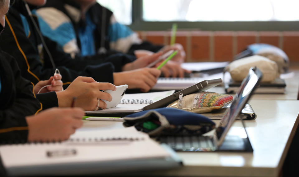 A student in uniform distracted holding using and watching a mobile phone during a lesson at high school. Books, tablets and pencil cases all visible on the desk and work space.