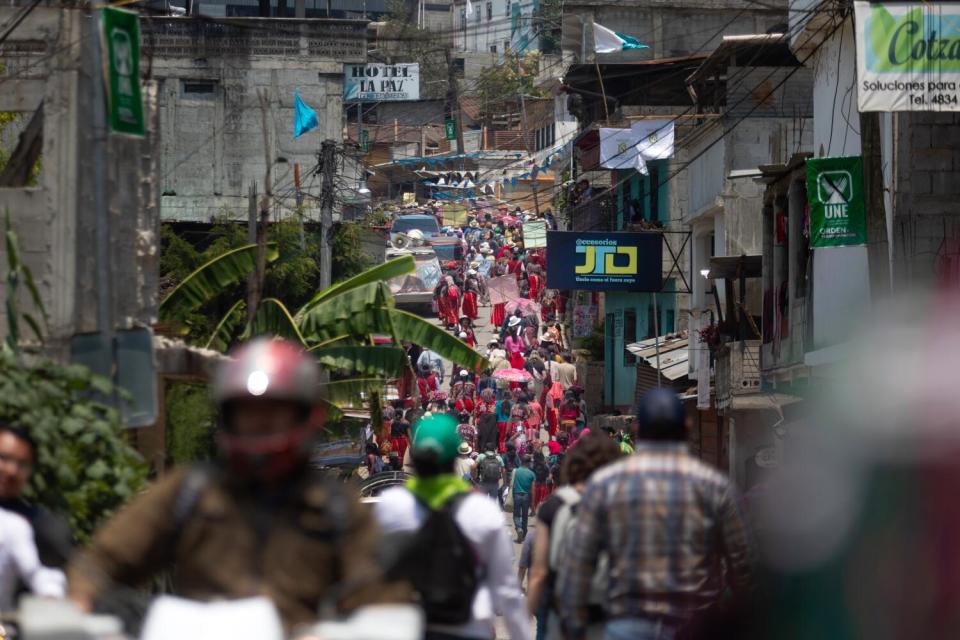 People in colorful red dress walk up a hill, flanked by buildings