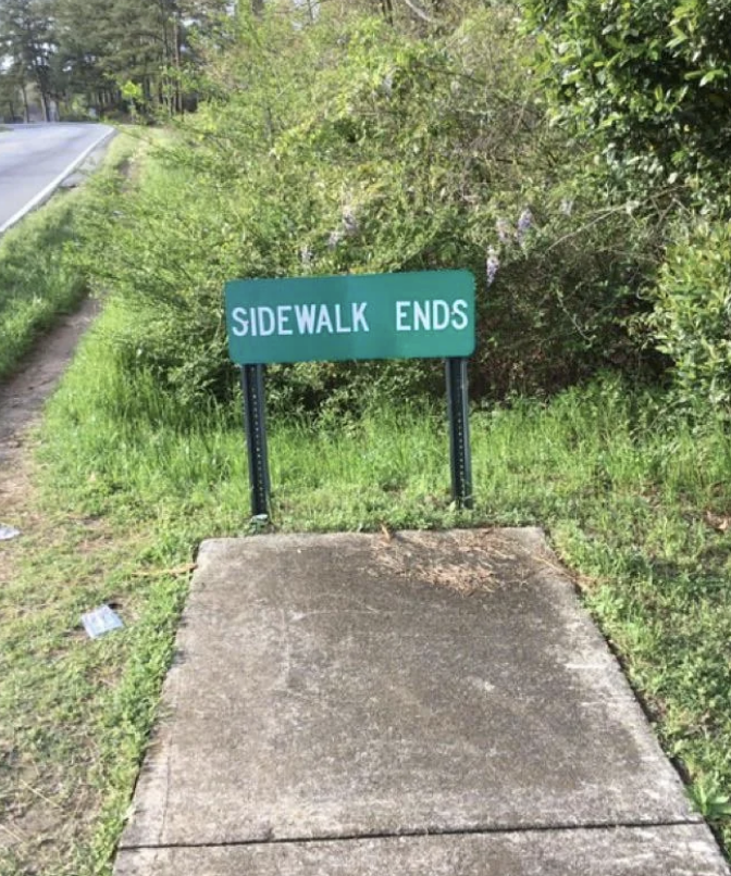 A sidewalk ending abruptly with a green sign that reads "SIDEWALK ENDS" in a grassy area beside a road