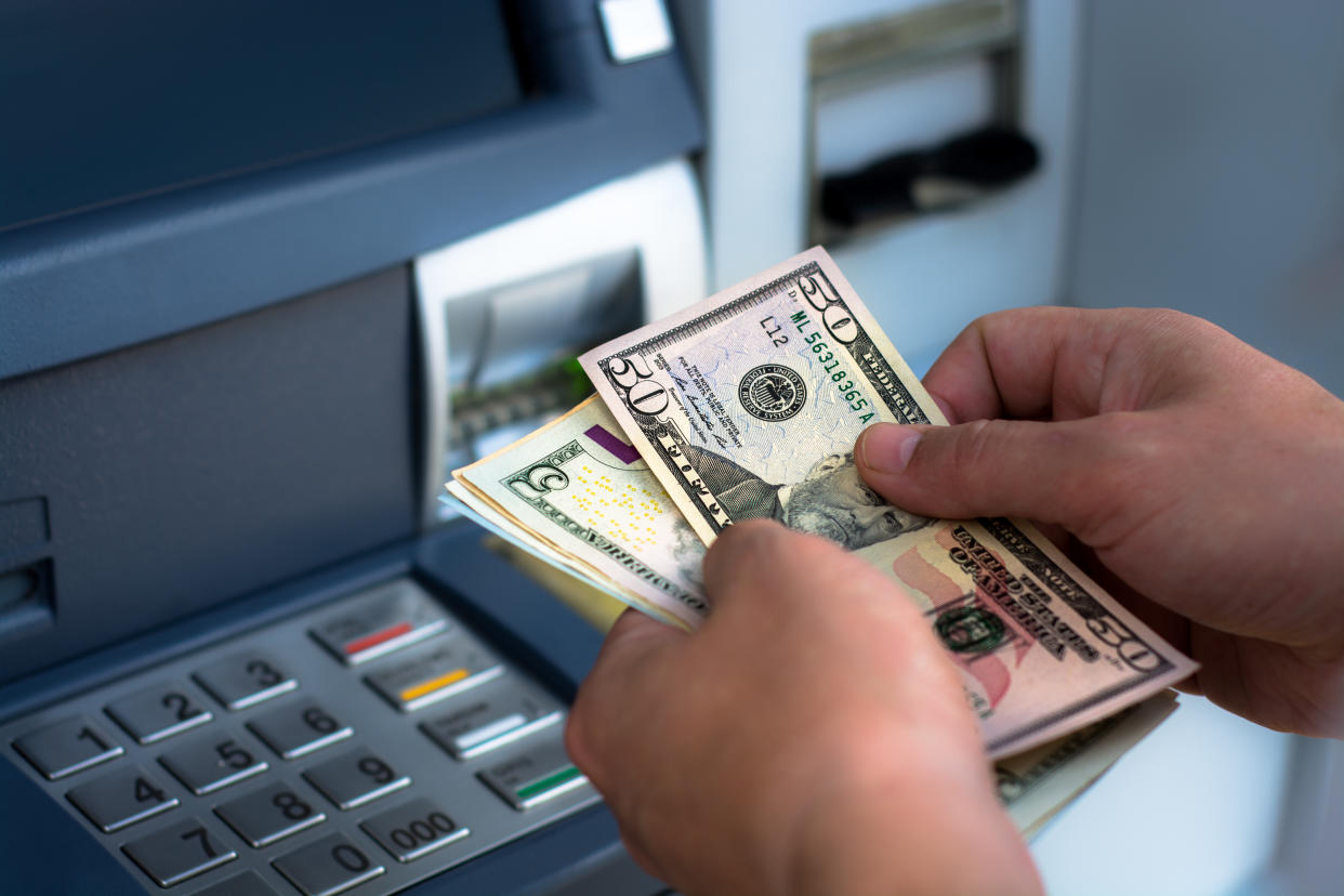 Hand are visible holding a stack of bills of U.S. currency in front of the keypad of an ATM.