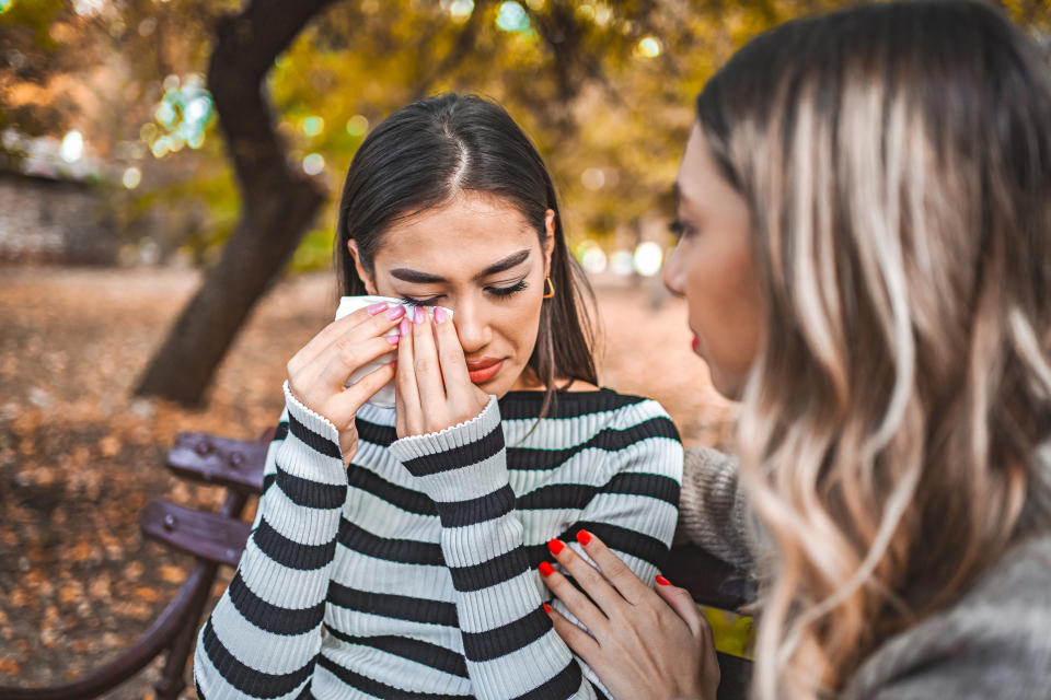 Two women are on a bench in a park. One woman cries, wiping her tears with a tissue, while the other comforts her with a hand on her shoulder