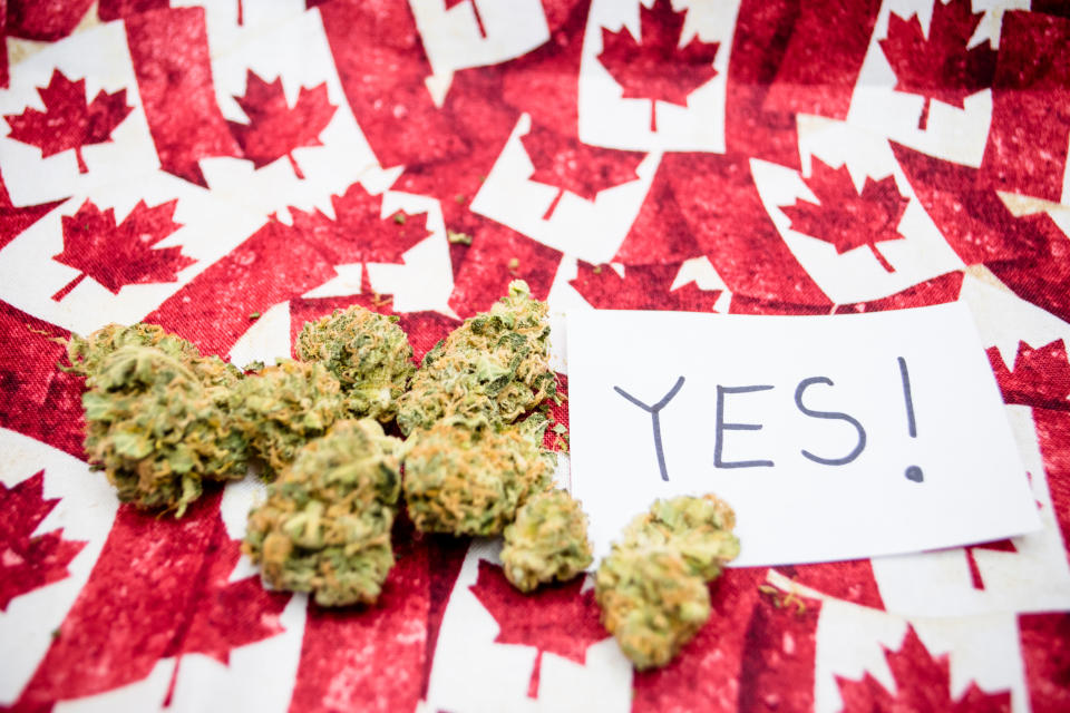 Dried cannabis buds next to an index card with the word "ye"s written on it, all lying atop dozens of miniature Canadian flags.