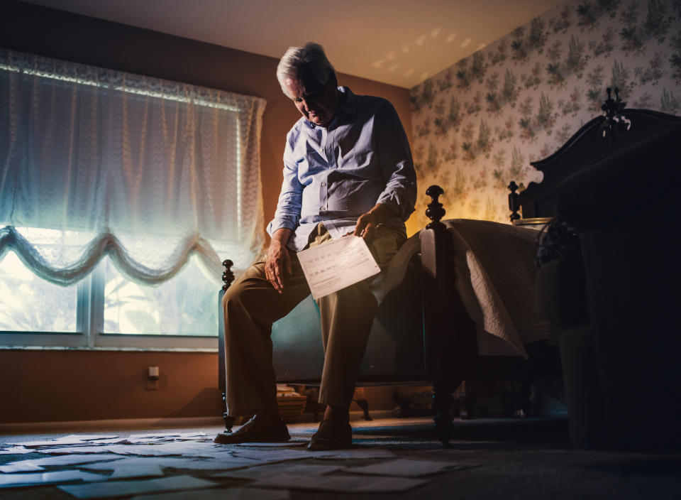 An elderly man sits on a bed, holding a sheet of paper, surrounded by scattered papers in a dimly lit room