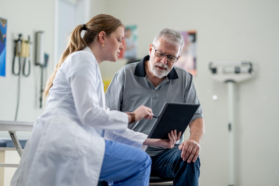 A senior gentleman meets with his female doctor to discuss his health concerns. He is dressed casually and seated in front of the doctor as she shares some recent test results with him on a tablet.