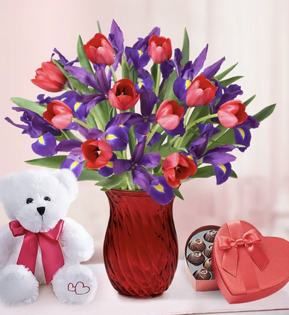 Red tulips and purple irises in a red vase surrounded by white teddy bear and heart shaped box of chocolates. 