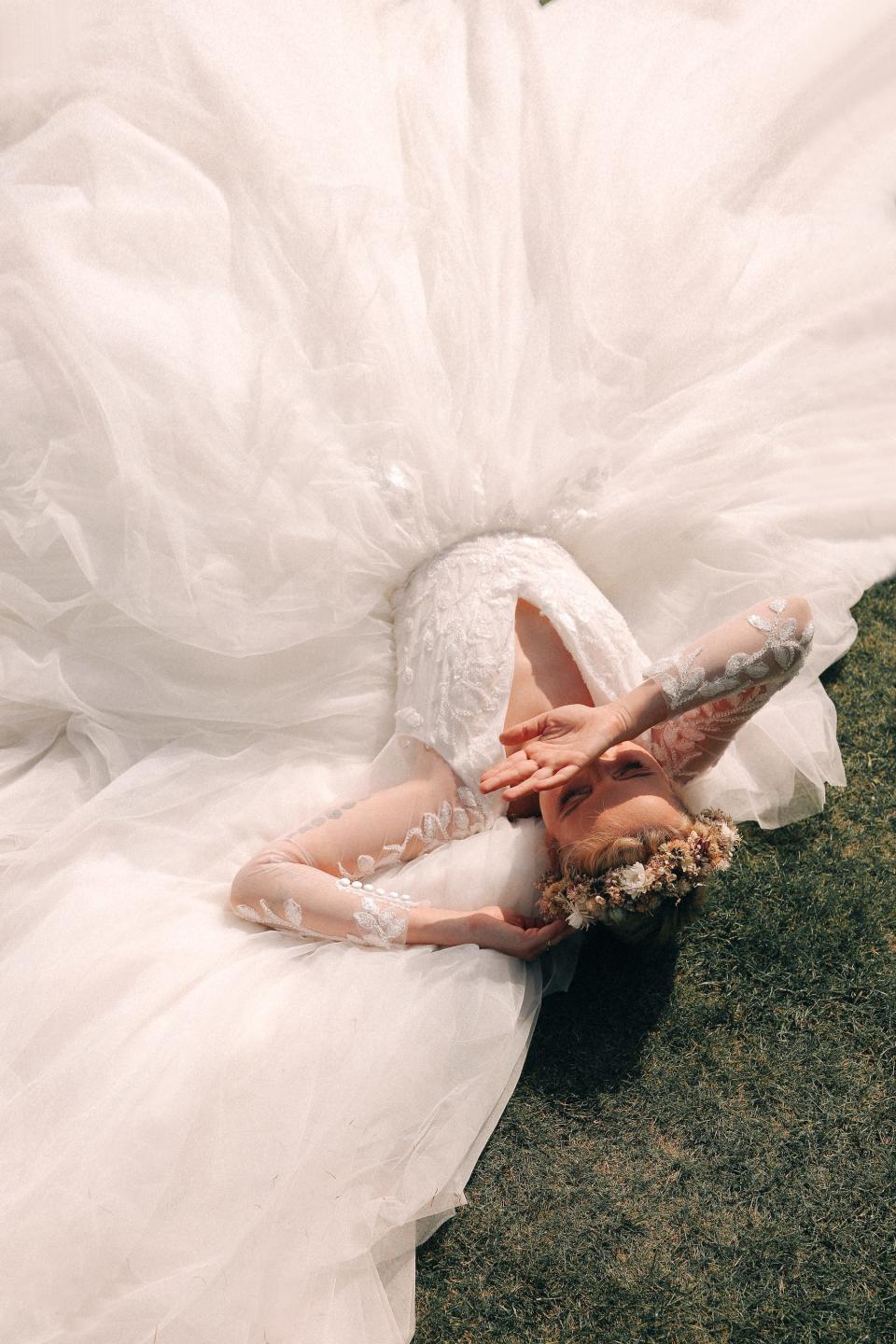 A person in a wedding dress lays on grass and covers her face.