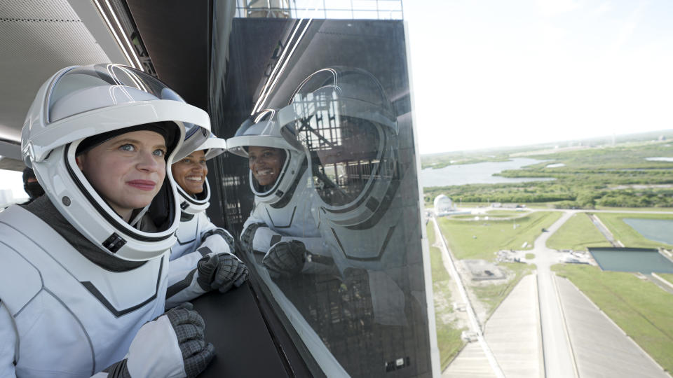 Two women in spacesuits look out a window showing a landscape with trees, water and roads